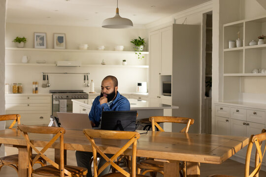 African man wearing denim shirt working on two laptops with cables at dining table in kitchen