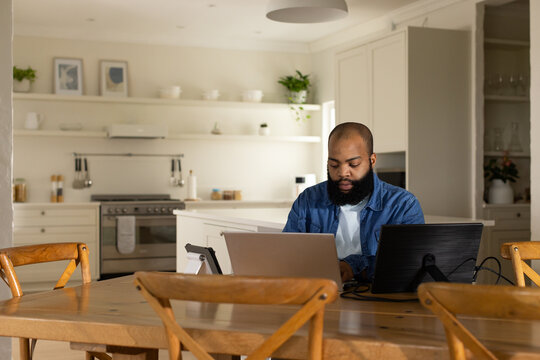 African American man working on laptop and monitor at table in kitchen in denim shirt
