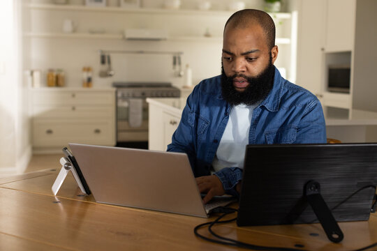 African American man sitting at wooden table in kitchen, working on laptop and tablet