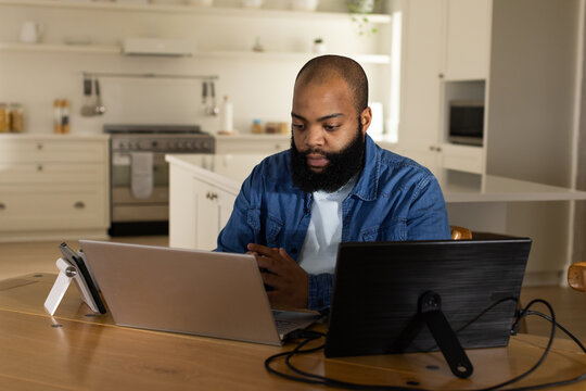 African American man wearing denim shirt sitting at table in kitchen, using laptop, tablet, phone