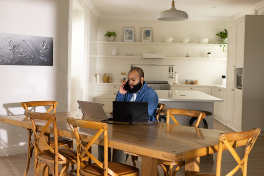 African American man in denim shirt working on laptops speaking on phone at wooden dining table