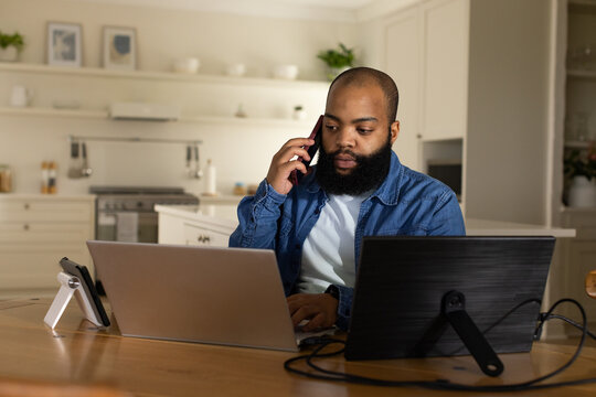Bearded man working in kitchen using dual screens and speaking on phone stand, in denim jacket