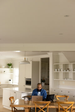 African American man wearing blue shirt sitting at wooden dining table working on laptop in kitchen