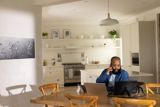 Adult African American man sitting at kitchen table calling on phone, using two laptops, copy space