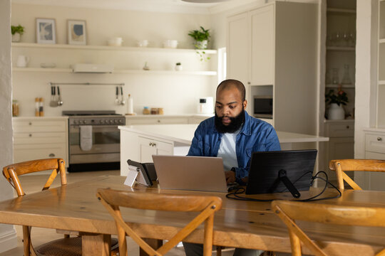 African American man sitting at table in kitchen, wearing denim shirt working on laptop, monitor