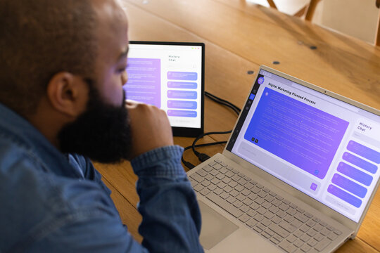 Bearded man sitting at wooden table working on laptop and external monitor with cables, copy space