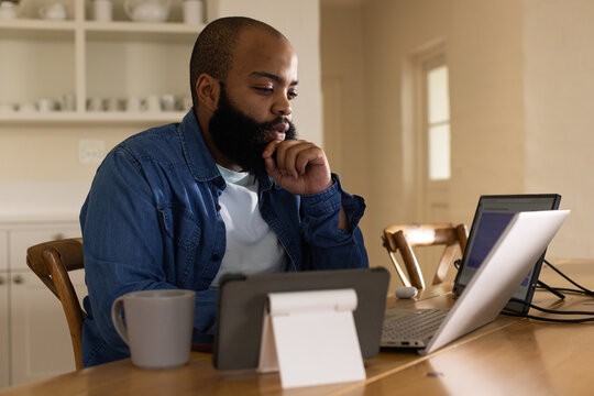 Adult African American man wearing denim shirt working on laptop with tablet, earbud case and mug