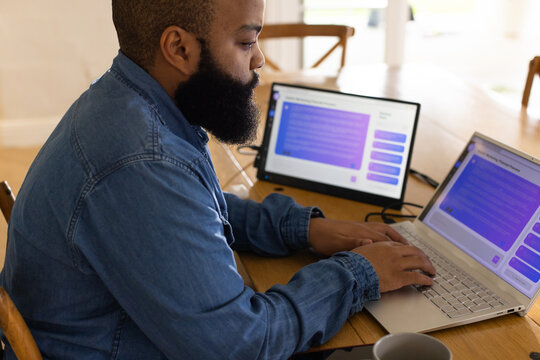 African man in denim shirt typing on laptop and checking tablet at dining table, copy space