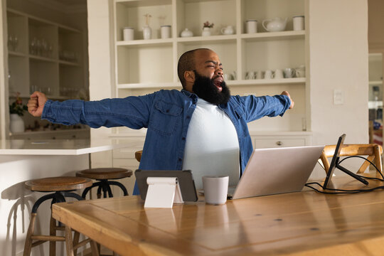 African American man stretching arms wide and yawning at wooden dining table workspace with laptop