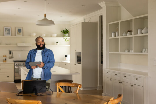 Adult African American man wearing denim shirt by kitchen island holding tablet working on laptop