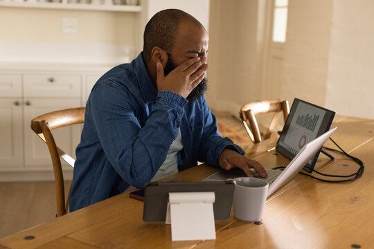 African American man in denim shirt rubbing eyes, working on laptop at kitchen table with monitor