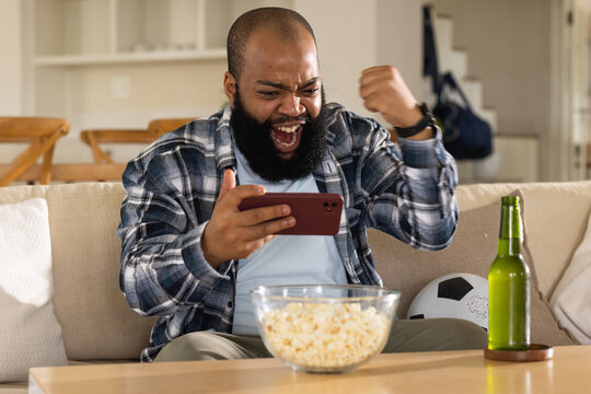 African American man sitting on couch at home holding red phone, cheering with popcorn and beer