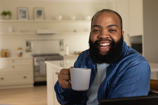 African American man sitting at kitchen island wearing denim shirt holding white mug and smiling