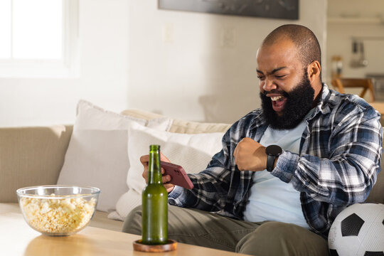 African American man reacting to smartphone on sofa in plaid shirt with popcorn and soccer ball