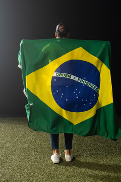 Adult female standing on studio turf holding Brazil flag draping back in blue jeans white sneakers
