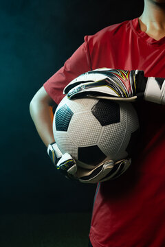 African American female goalkeeper holding soccer ball, wearing red jersey and gloves, showing turf