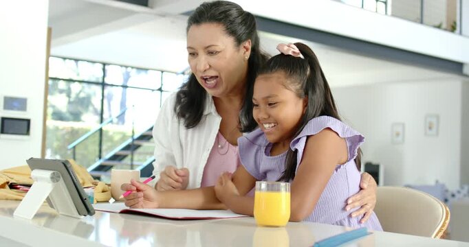 African American mom pointing tablet, guiding daughter writing notebook in kitchen with pink pen