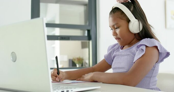 African American girl child following laptop lesson, writing notes at table wearing headphones