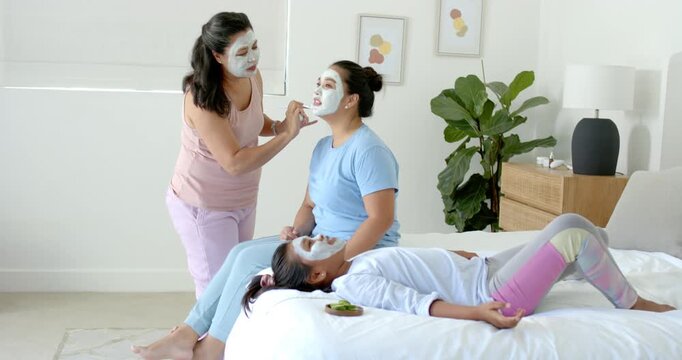 Diverse family mature woman applying mask with brush, green paste beside youth for pampering on bed
