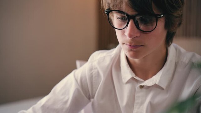Pensive teenage boy wearing eyeglasses and white shirt sitting at desk, deeply engrossed in studies and homework. Concept diligent and focused learning process