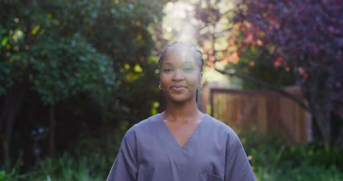Woman wearing gray scrub top tilting up, responding to vertical sunbeam, turning, smiling in garden