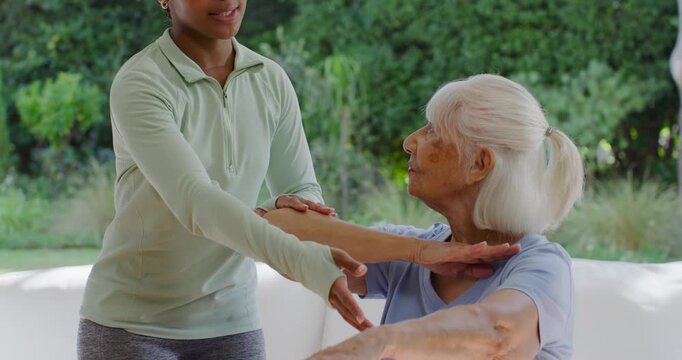 Adult African American caregiver showing arms, guiding senior raising arms for rehab on white sofa