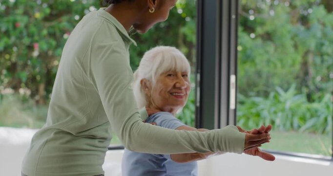 Diverse female caregiver placing hands on senior woman's arms guiding stretches by window for rehab