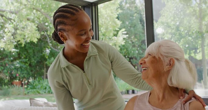 African American female carer in green holding senior in pink guiding exercise at home window