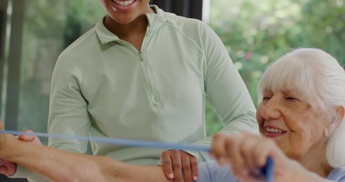 Diverse caregiver presenting blue resistance band and guiding senior in arm rows at home for rehab