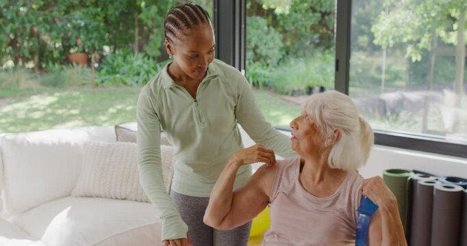 Diverse caregiver guiding senior woman in home for posture, leaning, pointing, senior lifting band