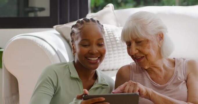 Diverse female helper and senior on couch younger sharing tablet content senior tapping laughing