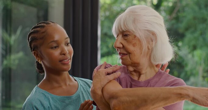 Female aide placing hands, guiding senior woman doing shoulder stretch near glass doors for rehab