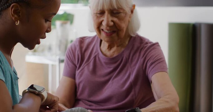 African American caregiver leaning in pointing at smartwatch  and  tapping phone guiding senior at h