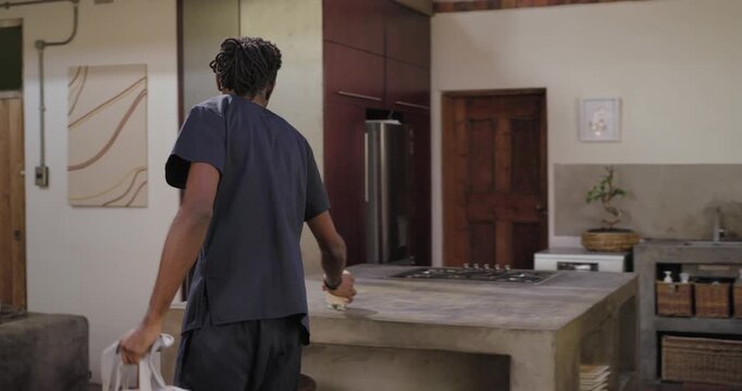 African adult man entering right, wiping island, opening bag, placing laptop, preparing for work