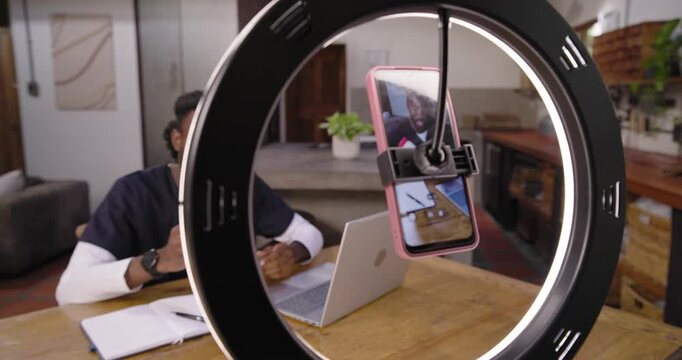 Adult African man using ring light replying to caller showing bottle writing notes at kitchen table