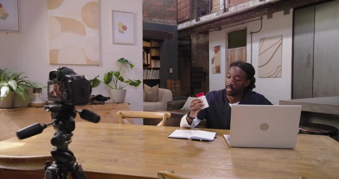 African American man in scrubs filming with DSLR at loft table checking notes explaining pill label