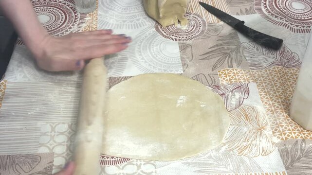 A woman rolls out raw dough with a wooden rolling pin on a patterned table, home kitchen setting. Homemade pastry preparation, traditional baking, culinary process.