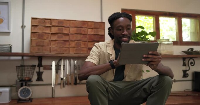African American man sitting on kitchen bench holding tablet, reacting to content while scrolling