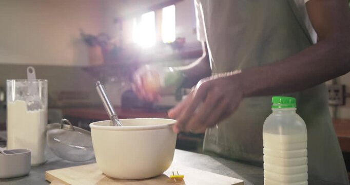 African man wearing apron cracking egg, steadying mixing bowl, whisking in kitchen preparing batter