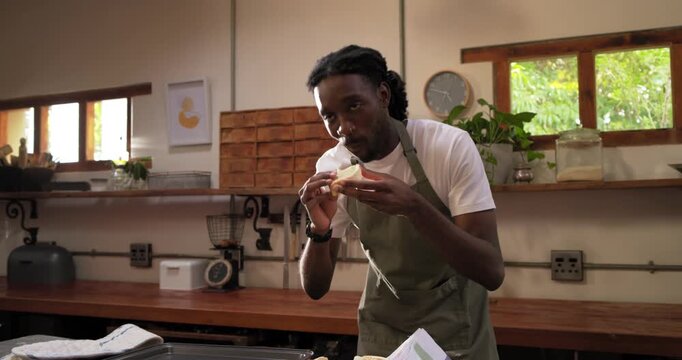 African American man in apron lifting roll on kitchen counter, tasting tearing and checking crumb
