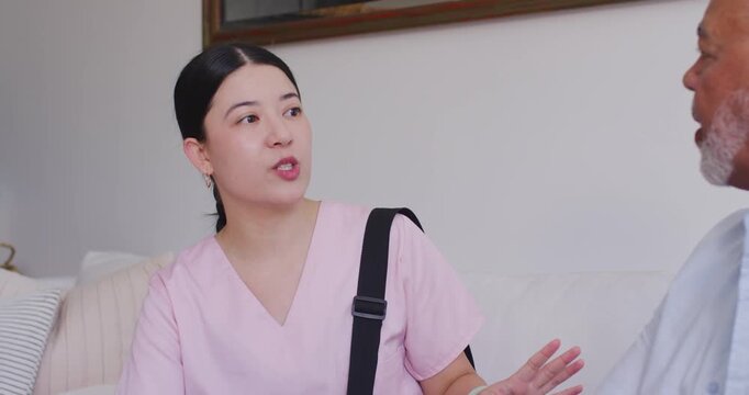Asian nurse in scrubs sitting sofa beside senior man, raising hand to clarify and reading notes