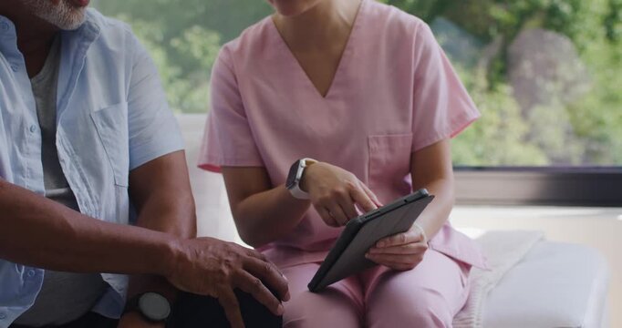 Asian nurse in pink scrubs tapping tablet and explaining care to African American patient on couch
