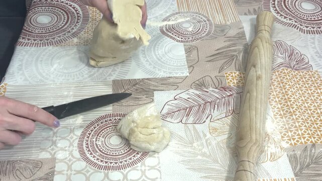 A woman cuts raw dough into large pieces with a knife on a patterned tablecloth, home kitchen setting. Homemade pastry preparation, traditional baking, culinary process.
