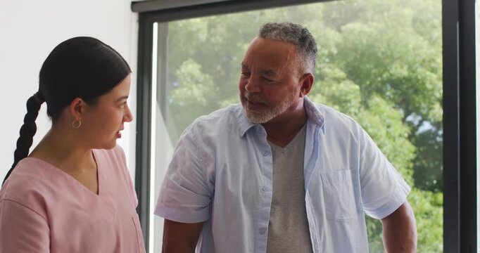 Asian caregiver in scrubs showing chart by window, client leaning nodding and agreeing plan