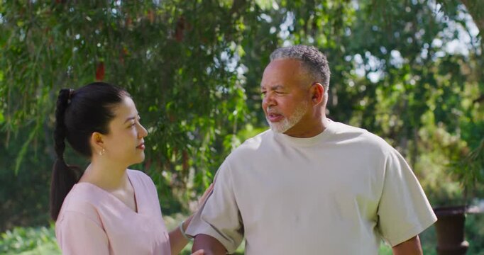 Asian nurse in pink scrubs steadying mature man showing uncertain look, guiding him across garden