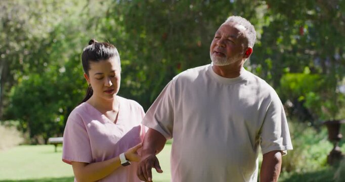 Asian caregiver in pink scrubs guiding senior man after watch alert steadying him walking in park