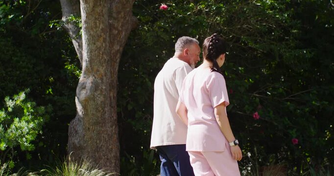 African American man and Asian aide in scrubs walking for health by tree, checking smartwatch