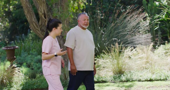 Asian female nurse in pink scrubs speaking, escorting African American senior man guiding in garden