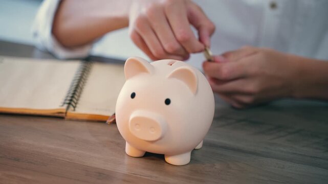 Close-up of person hand inserting coins into pink piggy bank on wooden table. Concept smart money management, financial savings, and investment for future security