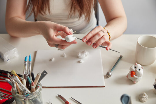 Close up of female hands sculpting a small animal figurine from white polymer clay on a white desk with professional tools.